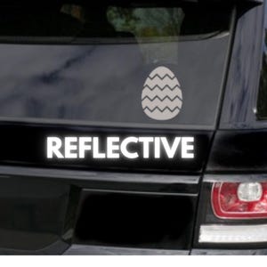Może przedstawiać: Odblaskowa naklejka na samochód z białym wzorem pisanki i zygzakowatym wzorem. Słowo "REFLECTIVE" jest wyświetlane pogrubionymi, białymi wielkimi literami pod jajkiem. Naklejka znajduje się na czarnym tyle pojazdu.
