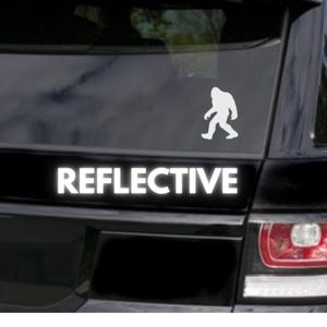 May include: A black car with a white silhouette of Bigfoot walking on the rear window. The car has a white reflective sticker that says "REFLECTIVE" on the rear bumper.