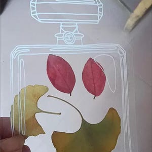 Könnte beinhalten: Eine durchsichtige Glasflasche mit weißem Umriss, die gepresste Blätter enthält. Die Blätter sind rot und gelb, und die Flasche hat ein weißes Chanel-Logo oben.