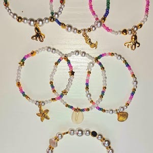 May include: Several beaded bracelets with pearl-like beads and colorful accents. Some bracelets feature gold charms, including an octopus, mermaid, starfish, and seashell. The bracelets are arranged on a white surface.
