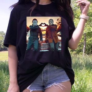 May include: Black t-shirt featuring a graphic of two horror movie characters forming a heart with their hands. The image shows Michael Myers and Jason Voorhees standing in front of a classic car. The shirt is a casual wear item.