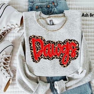 May include: Ash gray sweatshirt with the word "Dawgs" in red lettering over a leopard print design. The sweatshirt is paired with blue jeans and white sneakers. A gold chain necklace is visible.