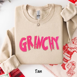 Puede incluir: Sudadera de color beige con la palabra "GRINCHY" en rosa brillante, contorneada en dorado. La sudadera tiene cuello redondo y mangas largas. Se ve un collar plateado con un colgante de sol. La palabra "Tan" est&aacute; impresa en la parte inferior. Art&iacute;culos navide&ntilde;os en el fondo.