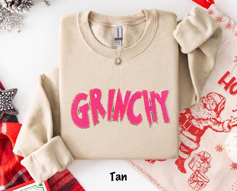 Puede incluir: Sudadera de color beige con la palabra "GRINCHY" en rosa brillante, contorneada en dorado. La sudadera tiene cuello redondo y mangas largas. Se ve un collar plateado con un colgante de sol. La palabra "Tan" est&aacute; impresa en la parte inferior. Art&iacute;culos navide&ntilde;os en el fondo.