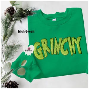 Irish green sweatshirt with the word "GRINCHY" embroidered in a lighter green, with a matching face mask. The sweatshirt is displayed on a white surface with pine and decorative elements.