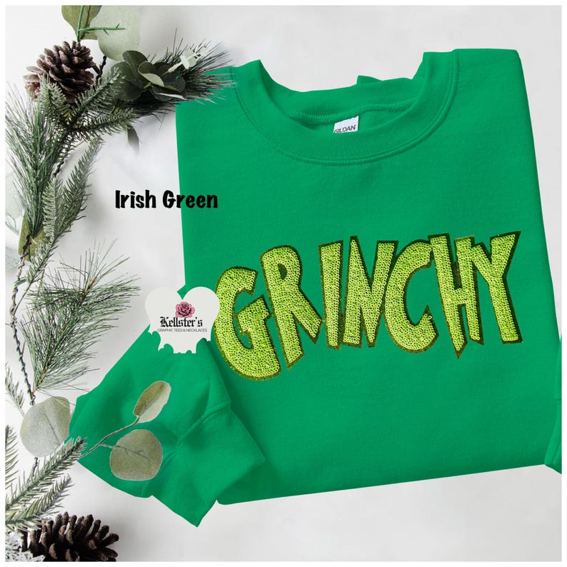 Irish green sweatshirt with the word "GRINCHY" embroidered in a lighter green, with a matching face mask. The sweatshirt is displayed on a white surface with pine and decorative elements.
