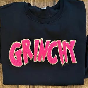 Puede incluir: Sudadera negra con la palabra "GRINCHY" en letras grandes, rosas y texturizadas con contorno dorado. La sudadera est&aacute; doblada sobre una superficie de madera. Una prenda casual y c&oacute;moda.
