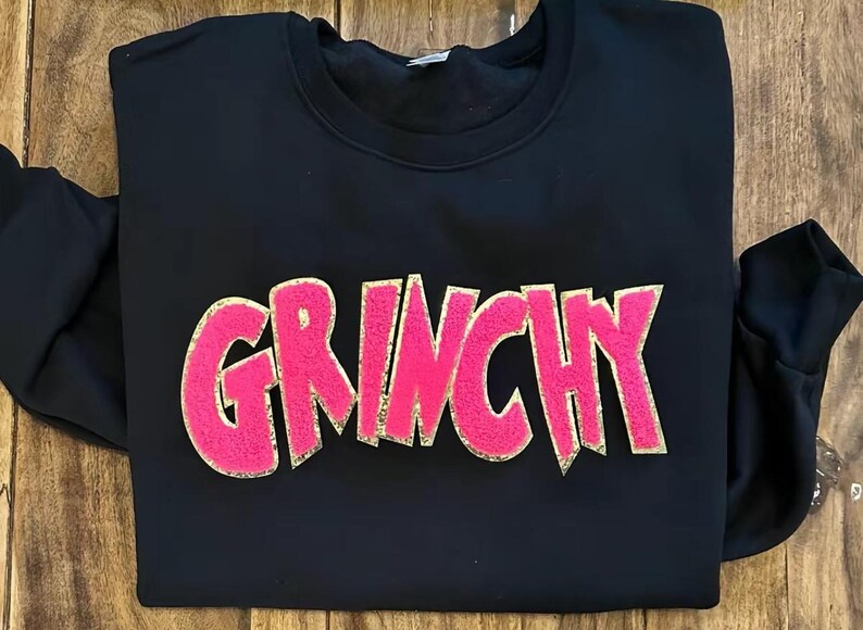 Puede incluir: Sudadera negra con la palabra "GRINCHY" en letras grandes, rosas y texturizadas con contorno dorado. La sudadera est&aacute; doblada sobre una superficie de madera. Una prenda casual y c&oacute;moda.