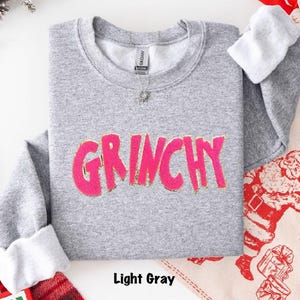 Puede incluir: Sudadera gris claro con la palabra "GRINCHY" en letras rosa brillante con ribete dorado. Las mangas est&aacute;n rematadas con tela blanca. El texto "Light Gray" est&aacute; en la parte inferior de la imagen.