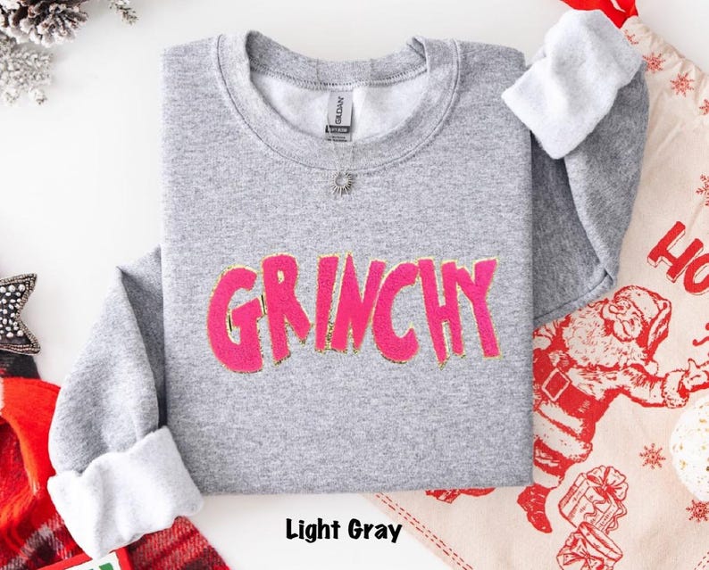 Puede incluir: Sudadera gris claro con la palabra "GRINCHY" en letras rosa brillante con ribete dorado. Las mangas est&aacute;n rematadas con tela blanca. El texto "Light Gray" est&aacute; en la parte inferior de la imagen.