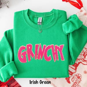 Puede incluir: Una sudadera verde con la palabra "GRINCHY" en rosa, con detalles de purpurina dorada. La sudadera tiene cuello redondo y mangas largas. Se ve un collar plateado con un colgante de sol. El texto "Irish Green" est&aacute; en la parte inferior.