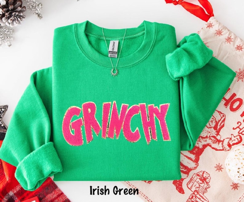 Puede incluir: Una sudadera verde con la palabra "GRINCHY" en rosa, con detalles de purpurina dorada. La sudadera tiene cuello redondo y mangas largas. Se ve un collar plateado con un colgante de sol. El texto "Irish Green" est&aacute; en la parte inferior.