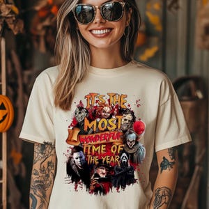 May include: Cream-colored t-shirt featuring a graphic with horror movie characters and the text "It's the Most Wonderful Time of the Year." The design includes images of iconic villains and a red banner.