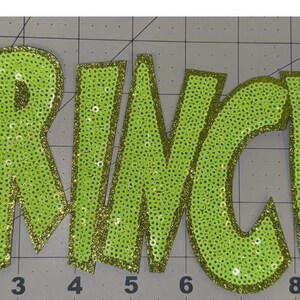 May include: Lime green sequined applique spelling out the word "GRINCHY". The letters are outlined in gold glitter and are on a gray cutting mat. The applique is likely for clothing or craft projects.