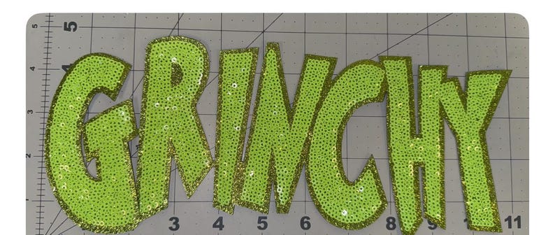 May include: Lime green sequined applique spelling out the word "GRINCHY". The letters are outlined in gold glitter and are on a gray cutting mat. The applique is likely for clothing or craft projects.