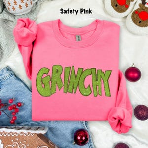 pink sweatshirt with the word "GRINCHY" in green sequined letters. The text "Safety Pink" is above the sweatshirt. The sweatshirt is surrounded by Christmas decorations, including ornaments and a gingerbread house.
