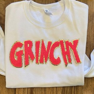 Puede incluir: Camiseta blanca de manga larga con la palabra "GRINCHY" en rojo brillante, contorneada con purpurina dorada. La camiseta est&aacute; hecha de un material suave. La camiseta est&aacute; colocada sobre una superficie de madera.