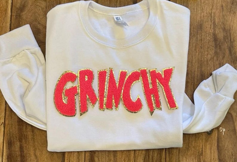 Puede incluir: Camiseta blanca de manga larga con la palabra "GRINCHY" en rojo brillante, contorneada con purpurina dorada. La camiseta est&aacute; hecha de un material suave. La camiseta est&aacute; colocada sobre una superficie de madera.