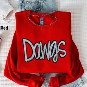 May include: A bright red long-sleeve crewneck sweatshirt featuring the word Dawgs in large, silver sequined lettering across the chest. The garment is displayed with a silver chain necklace resting on the collar and blue denim fabric visible.