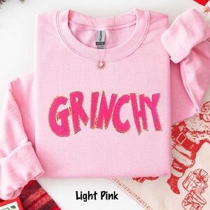 Puede incluir: Sudadera rosa claro con la palabra "GRINCHY" en rosa brillante, contorneada en dorado. La parte superior de manga larga est&aacute; doblada y se ve un collar peque&ntilde;o. Las palabras "Light Pink" est&aacute;n en la parte inferior.