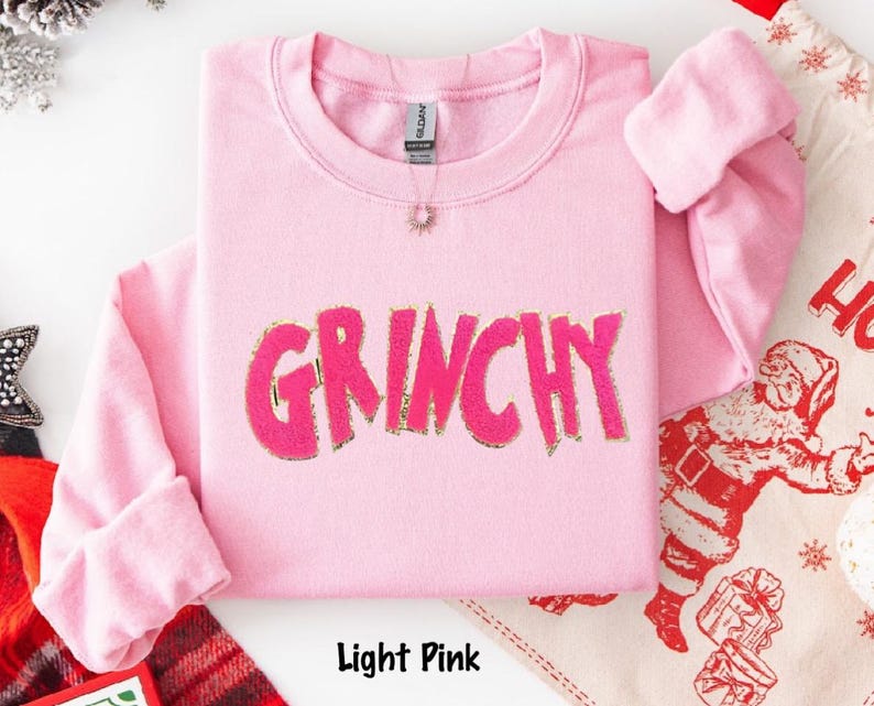 Puede incluir: Sudadera rosa claro con la palabra "GRINCHY" en rosa brillante, contorneada en dorado. La parte superior de manga larga est&aacute; doblada y se ve un collar peque&ntilde;o. Las palabras "Light Pink" est&aacute;n en la parte inferior.