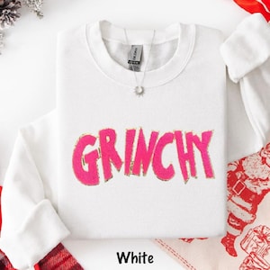 Puede incluir: Sudadera blanca con la palabra "GRINCHY" en rosa brillante, contorneada en dorado. La sudadera tiene cuello redondo y mangas largas. La palabra "White" est&aacute; impresa en la parte inferior de la imagen.