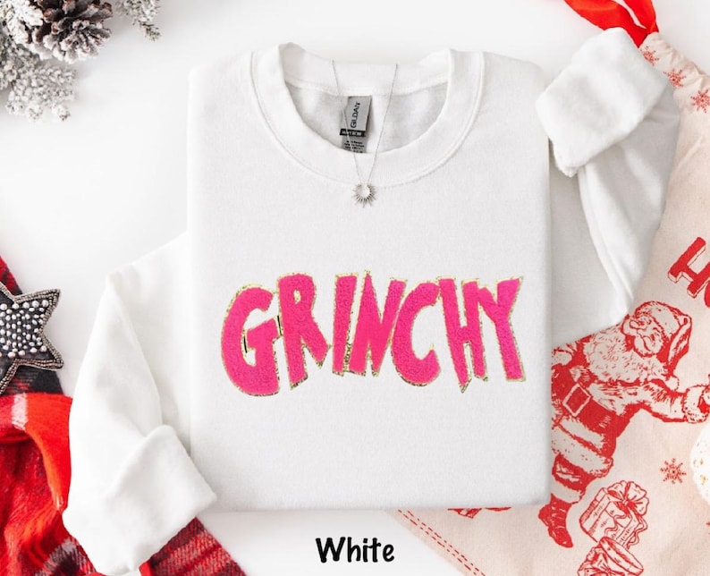 Puede incluir: Sudadera blanca con la palabra "GRINCHY" en rosa brillante, contorneada en dorado. La sudadera tiene cuello redondo y mangas largas. La palabra "White" est&aacute; impresa en la parte inferior de la imagen.