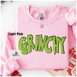 Light pink sweatshirt with the word "GRINCHY" in large, lime green, embroidered letters. The sweatshirt has a crew neck and long sleeves. The brand tag is visible at the back of the neck.