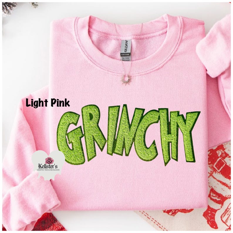 Light pink sweatshirt with the word "GRINCHY" in large, lime green, embroidered letters. The sweatshirt has a crew neck and long sleeves. The brand tag is visible at the back of the neck.