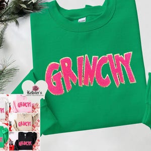 Puede incluir: Una sudadera verde con la palabra "Grinchy" en letras rosas y brillantes. La sudadera se muestra con otras opciones de color: blanco, rosa, beige, negro y verde. La sudadera es de Kellster's Graphic Tees & Necklaces.