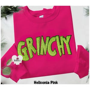 A Heliconia Pink sweatshirt with the word "GRINCHY" embroidered in lime green. The sweatshirt has a crew neck and long sleeves. The brand name "Kellster's" is visible on a small heart-shaped logo.n