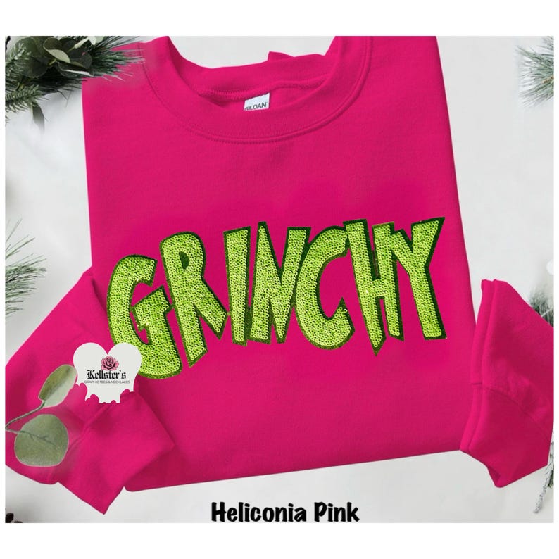 A Heliconia Pink sweatshirt with the word "GRINCHY" embroidered in lime green. The sweatshirt has a crew neck and long sleeves. The brand name "Kellster's" is visible on a small heart-shaped logo.n