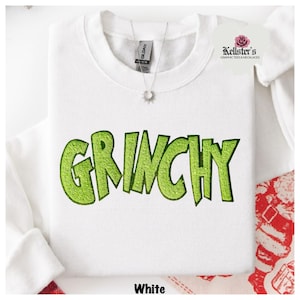 White sweatshirt with the word "GRINCHY" in bright green, sequined letters. A silver necklace with a sunburst pendant hangs near the collar.