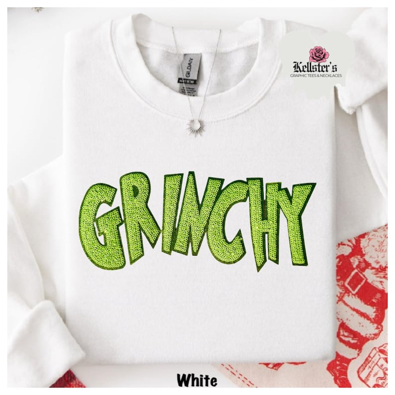 White sweatshirt with the word "GRINCHY" in bright green, sequined letters. A silver necklace with a sunburst pendant hangs near the collar.