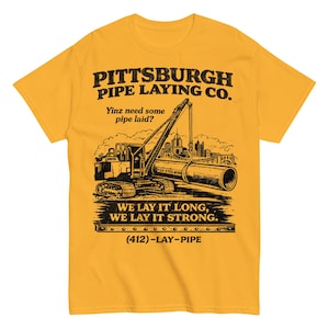 Könnte beinhalten: Senfgelbes T-Shirt mit einer schwarzen Grafik eines Baufahrzeugs, das ein Rohr anhebt, und dem Text "PITTSBURGH PIPE LAYING CO." und "WE LAY IT LONG, WE LAY IT STRONG." Das Shirt enthält auch den Text "Yinz need some pipe laid?" und die Telefonnummer (412)-LAY-PIPE.