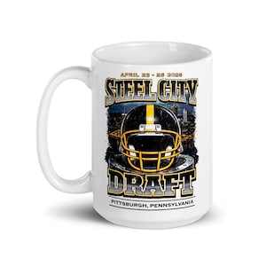 May include: White ceramic mug featuring a football helmet design with the text "STEEL CITY DRAFT" and "APRIL 23-25 2026". The mug also includes the city name "PITTSBURGH, PENNSYLVANIA". The design is in black, yellow, and gray.