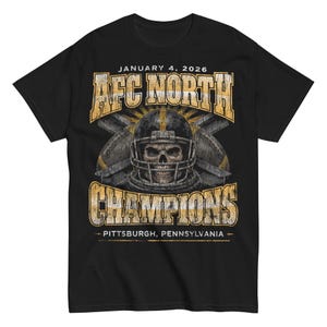 May include: Black t-shirt featuring a distressed graphic with the text "AFC North Champions" and "January 4, 2026". The design includes a football helmet with a skull and crossed footballs, with the location "Pittsburgh, Pennsylvania".