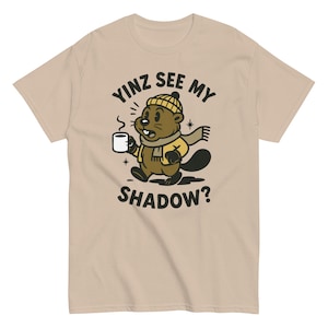 Könnte beinhalten: Beigefarbenes T-Shirt mit einem Cartoon-Biber, der eine Kaffeetasse hält. Der Biber trägt eine gelbe Jacke, eine Strickmütze und einen Schal. Der Text auf dem Shirt lautet "Yinz See My Shadow?" in einem kreisförmigen Design.