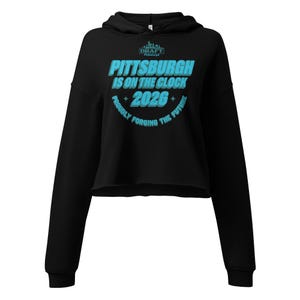 Puede incluir: Sudadera corta negra con capucha y mangas largas. En la parte delantera, el texto turquesa "PITTSBURGH IS ON THE CLOCK 2026", con "PROUDLY FORGING THE FUTURE" en un diseño circular. La sudadera tiene un cordón.
