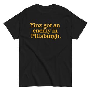 May include: Black t-shirt featuring the text "Yinz got an enemy in Pittsburgh" in gold. The shirt has a crew neck and is likely made from a soft fabric, suitable for casual wear.