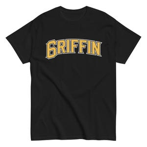 Puede incluir: Camiseta negra con la palabra "GRIFFIN" en dorado, con una fuente de estilo universitario con contorno blanco. La camiseta es de cuello redondo y manga corta.