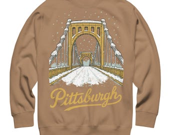 May include: A tan long-sleeve sweatshirt featuring a graphic illustration of a yellow suspension bridge in the snow. The bridge is depicted with white snow on its structure and the road surface. The word Pittsburgh is printed in yellow script below.