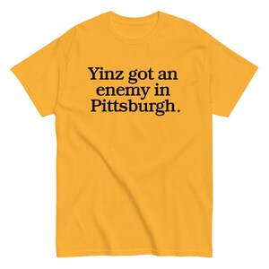 May include: Mustard yellow t-shirt with the black text "Yinz got an enemy in Pittsburgh". The shirt has a crew neck and short sleeves.