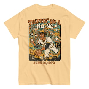 May include: A light yellow t-shirt featuring a retro graphic of a baseball player in a Pirates uniform. The design includes the text "Trippin' on a No-No" and "June 12, 1970". The illustration has a vintage, psychedelic style.