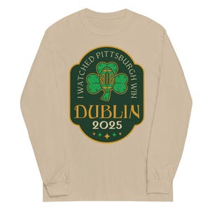 May include: A long-sleeve, sand-colored shirt featuring a green and gold graphic. The graphic includes a football-shaped shamrock with the text "I WATCHED PITTSBURGH WIN DUBLIN 2025". The design also has four green stars.