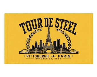 May include: A rectangular yellow towel featuring a black graphic print. The text reads TOUR DE STEEL, PITTSBURGH, PARIS, and OCTOBER 25, 2026. The design includes a central Eiffel Tower, a city skyline, laurel branches, and a small airplane icon.