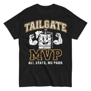 Peut inclure: T-shirt noir avec un motif blanc et jaune. Le motif présente les mots "TAILGATE MVP" au-dessus d'un tonneau de dessin animé avec des bras fléchissant. En dessous, le texte indique "ALL STATS, NO PADS."