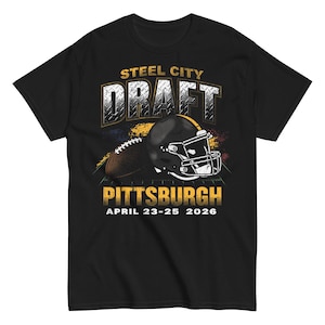 Pode incluir: Camiseta preta com um design de futebol americano e capacete. O texto diz "STEEL CITY DRAFT PITTSBURGH APRIL 23-25 2026" em ouro e branco. O design apresenta uma bola de futebol americano e um capacete preto.