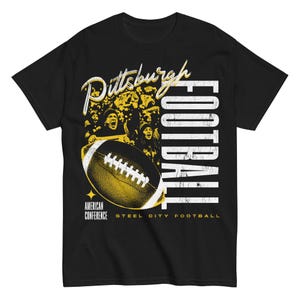 May include: Black t-shirt featuring a football design. The graphic includes the words "Pittsburgh Football", a football, and the text "American Conference Steel City Football". The design is in yellow and white.