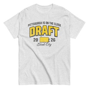Puede incluir: Camiseta gris claro con un gráfico amarillo y negro desgastado. El gráfico dice "PITTSBURGH IS ON THE CLOCK DRAFT 20 26 Steel City" e incluye un contorno amarillo de Pensilvania.